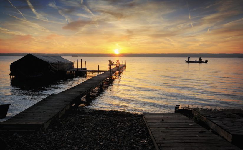 A beautiful autumn sunrise on the pebbled shores of Lake Cayuga in the Finger lakes region of New York state. A row boat with oars is docked on the side of a peir that leads out to a power boat shelter and a deck with chairs for watching the sunrise. Two fisherman enjoy the sunrise from their boat.